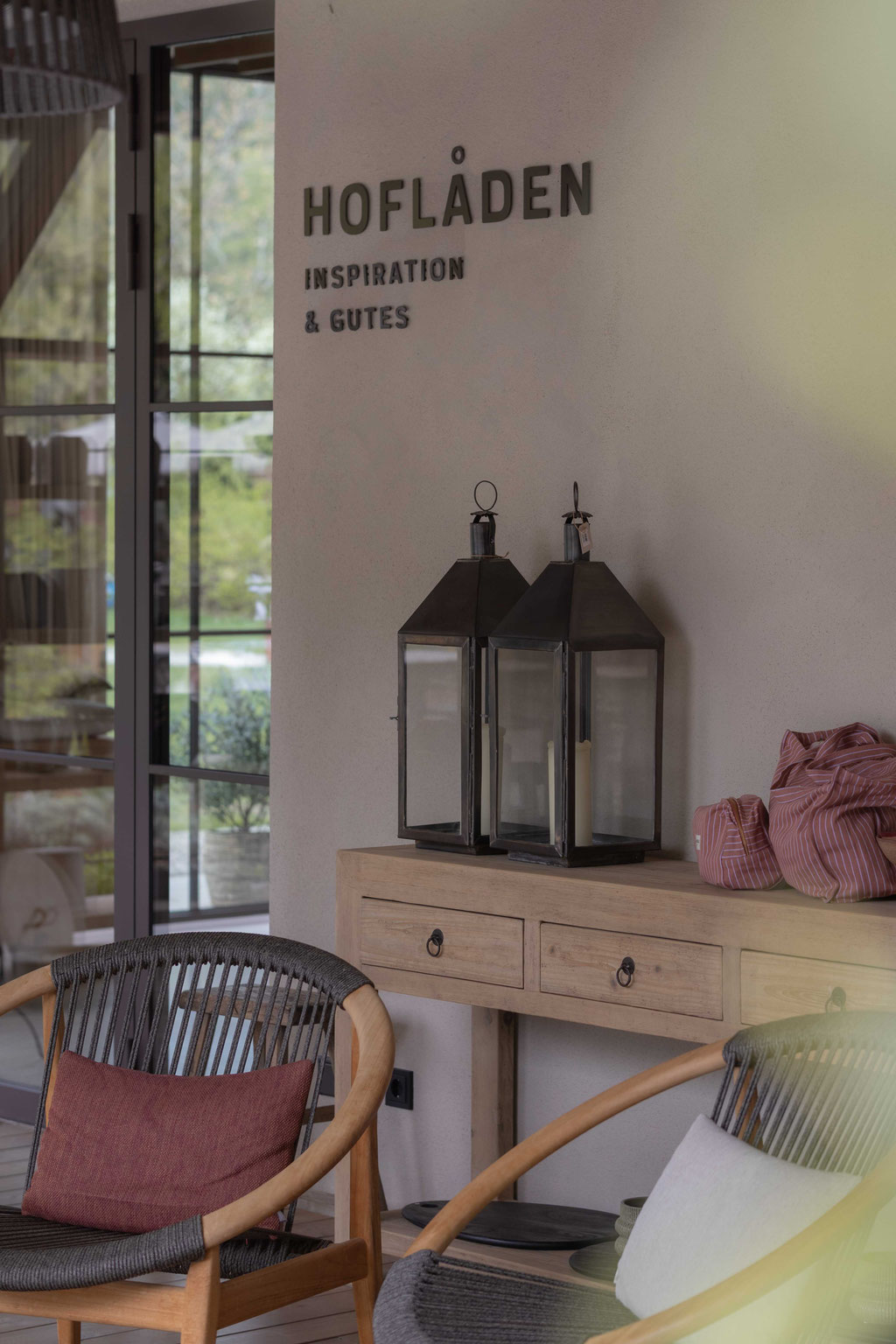 A cosy interior featuring a wooden console table, lanterns and armchairs, set beneath a wall sign reading “HOFLÅDEN – Inspiration & Good Things” next to large windows.