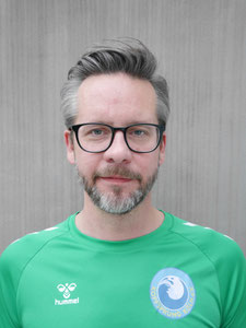 Florian: Mann mit Vollbart und markanter Brille, Seitenscheitel, leicht ergraut, schmunzelnd