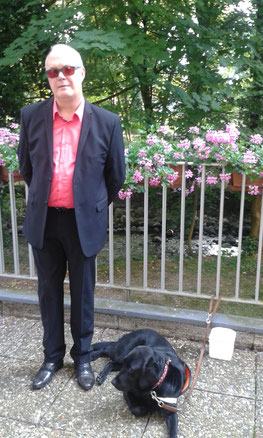 Philippe est habillé en costard et Jumpy est allongée à ses pieds devant une barrière qui donne un jardin en fleurs.