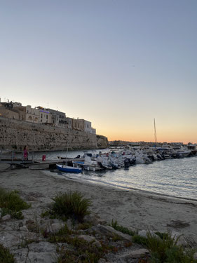 spiaggia con barchette attraccate al tramonto, un po' di fine sabbia bianca e in alto le mura della città di Otranto