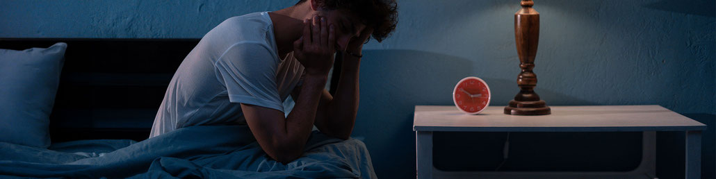 Mann sitzt am Bett, weil er nicht schlafen kann, es ist Nacht und der Wecker zeigt kurz vor drei