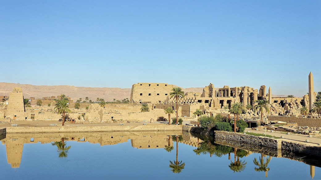 Ancient Egyptian temple ruins reflected in a still water basin, with palm trees and desert hills under a clear blue sky.
