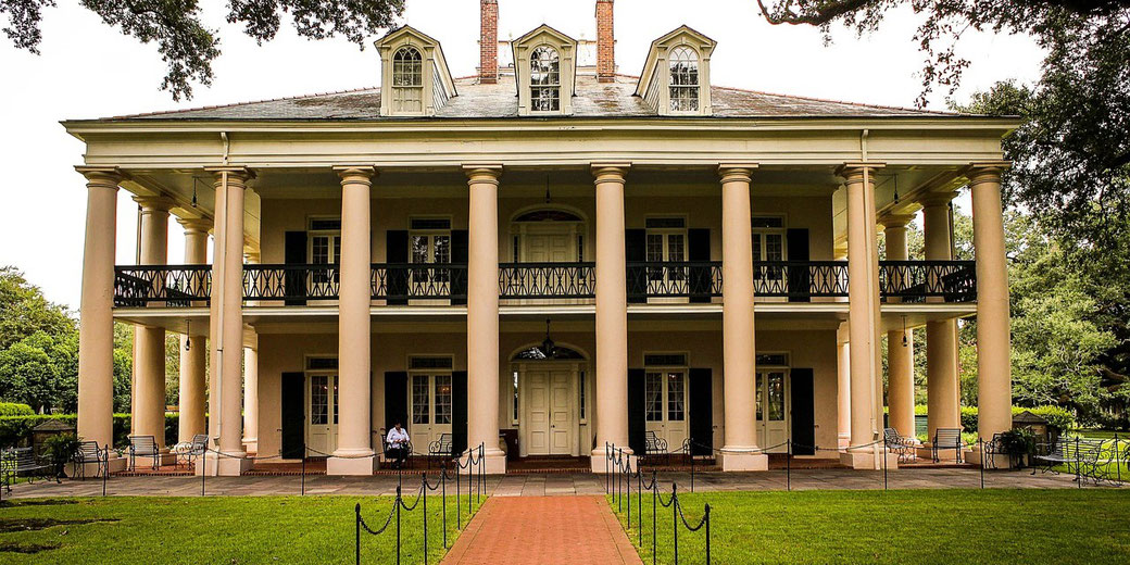 Large antebellum-style plantation house with tall columns, symmetrical design, and wraparound balconies, surrounded by trees and manicured lawn.