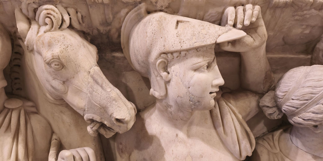 Marble relief of a soldier lifting his helmet beside a horse, with another figure partially visible to the right.