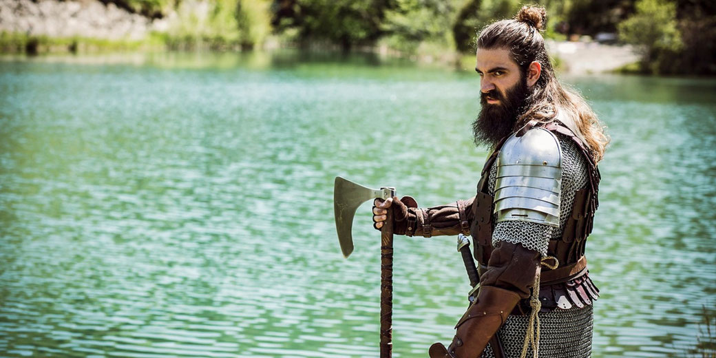 A bearded warrior in chainmail and plate armor stands by a lake, gripping a battle axe.
