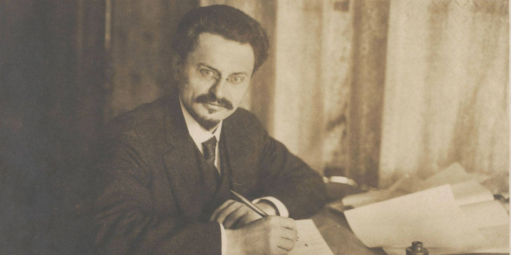 A man with glasses and a goatee sits at a desk, writing with a pen, surrounded by papers in a sepia-toned vintage photograph.