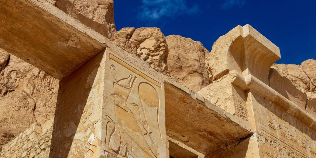 Ancient sandstone temple wall with carved hieroglyphs and a figure in profile, set against rugged desert cliffs under a clear deep blue sky.