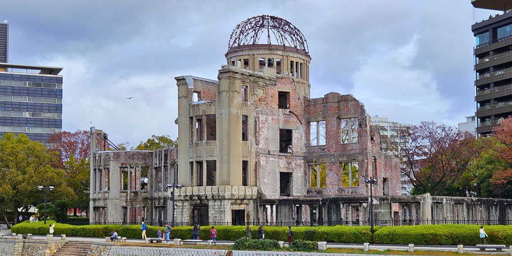The Atomic Bomb Dome in Hiroshima stands partially ruined, preserved as a memorial with visitors walking nearby and modern buildings in the background.