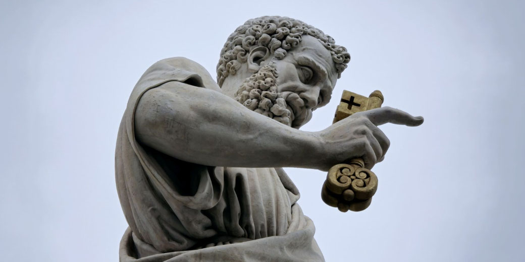 A close-up of a statue of St. Peter at the Vatican, holding a large golden key with a cross. His detailed beard, curly hair, and solemn expression stand out against the cloudy sky.