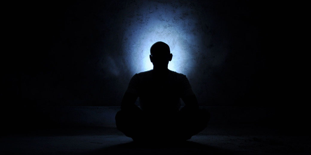 A person sits cross-legged in the dark with a faint blue glow illuminating the wall behind their silhouette.