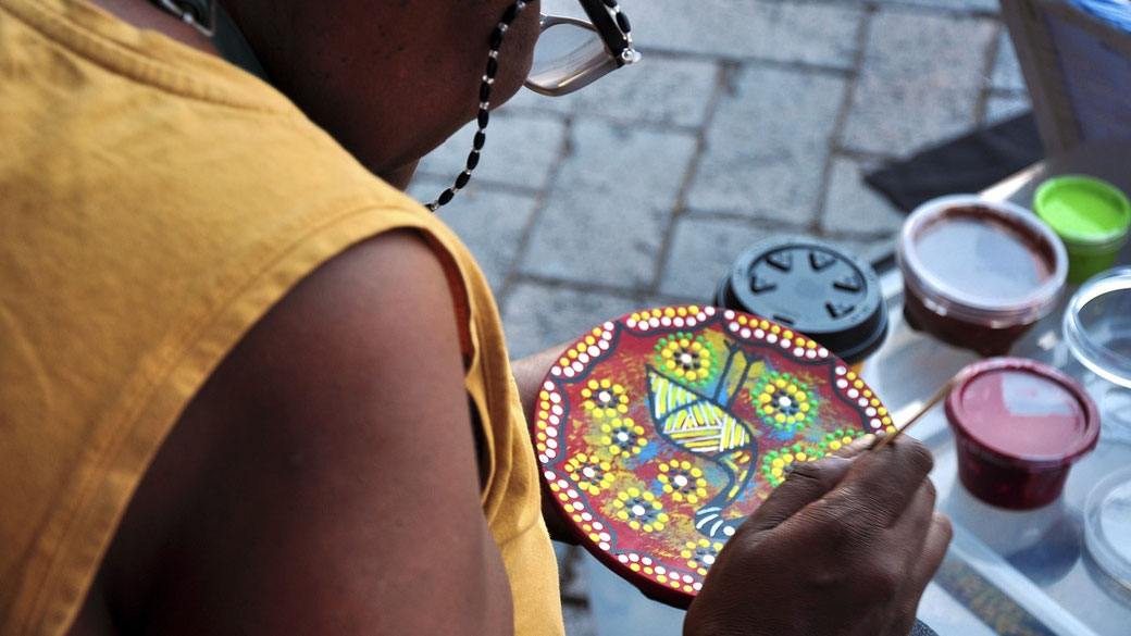 An artist in a yellow top paints a small circular plate with intricate Indigenous dot art, featuring a turtle design.