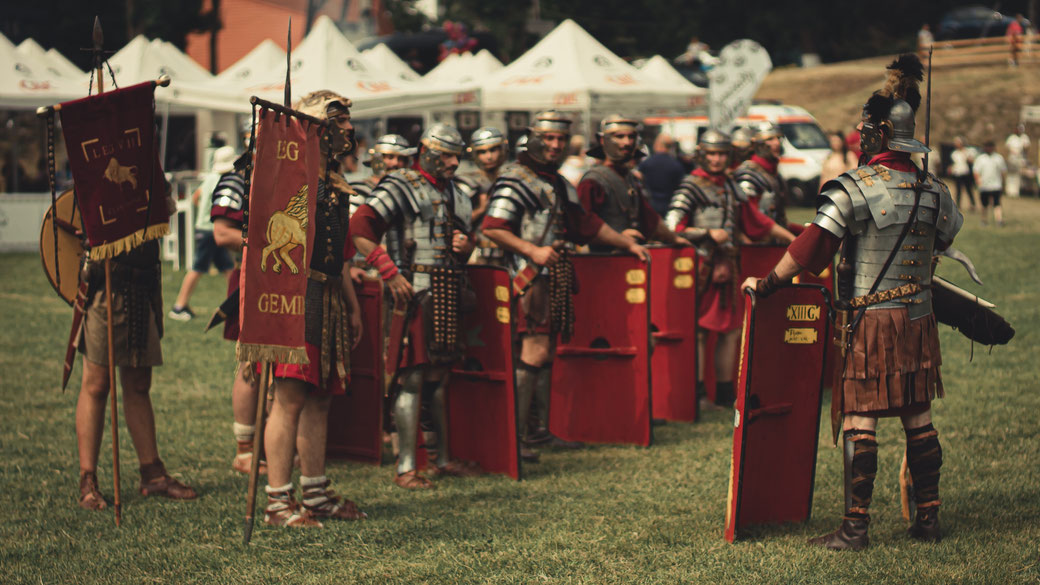 A group of reenactors dressed as Roman legionaries stand in formation with red shields and military standards, appearing to engage in a drill or ceremony at an outdoor historical event.
