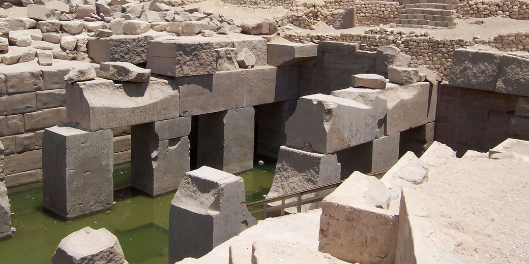 Ruins of an ancient Egyptian structure with massive stone blocks and standing pillars partially submerged in greenish water.