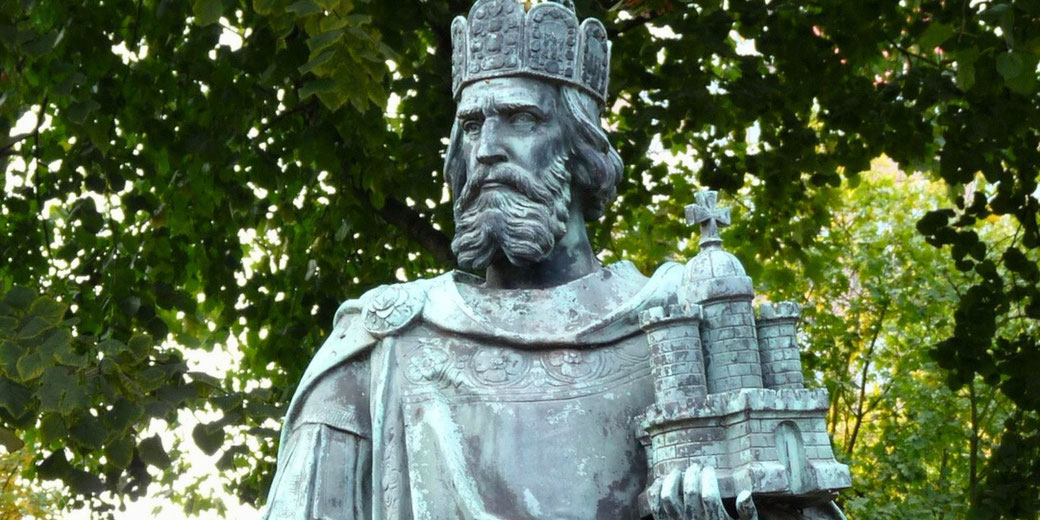 Bronze statue of a crowned king with a beard, holding a miniature model of a castle, surrounded by green leafy trees.