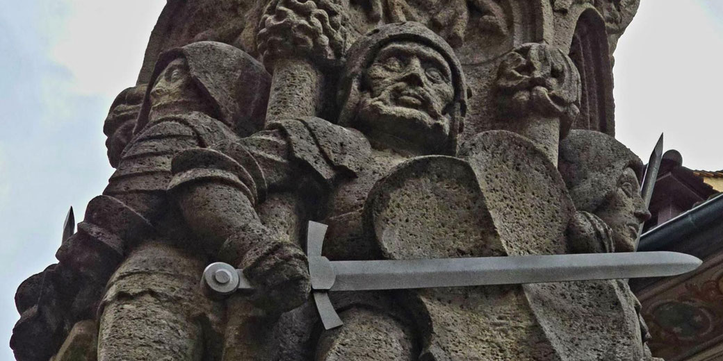 A stone statue of a medieval soldier holding a sword and shield, wearing armor and a helmet. The detailed carving emphasizes the warrior's stern expression and historical attire.