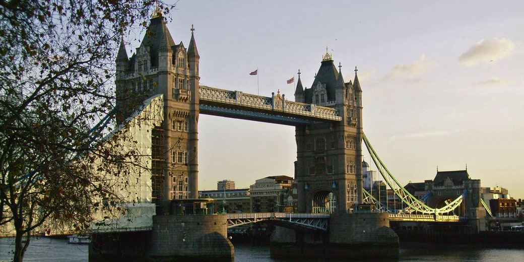 Tower Bridge in London is shown at sunset, with its twin towers and suspended walkways highlighted by golden light.
