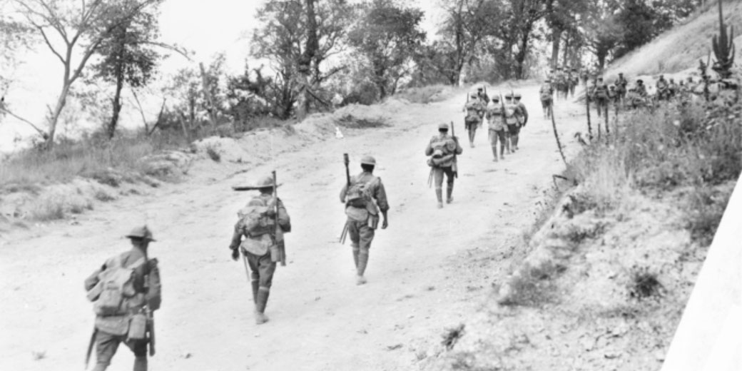 Soldiers equipped with gear and rifles march uphill on a dirt path surrounded by trees and sparse vegetation.