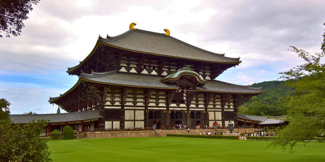 Tōdai-ji, a historic Buddhist temple in Nara, Japan. Its massive wooden hall features intricate architecture, a sweeping roof, and golden ornaments, surrounded by lush greenery and visitors.
