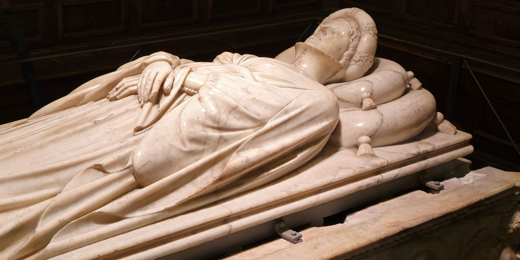 Marble effigy of a reclining figure lies atop a tomb, hands folded, head resting on tassel-trimmed cushions.