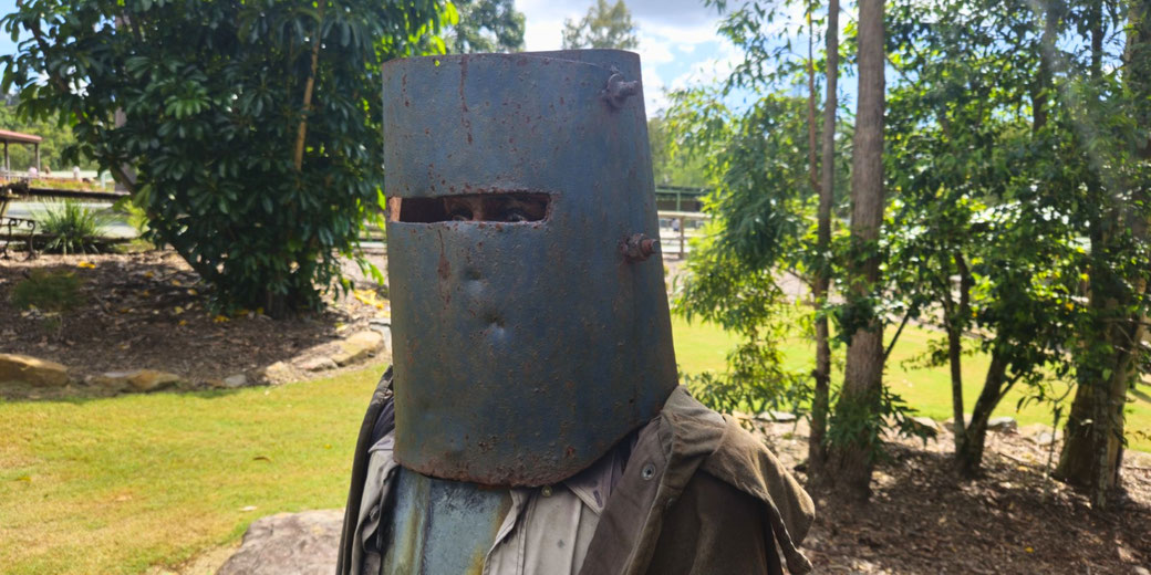 A person wears a large, rusted metal helmet with a narrow eye slit, evoking the appearance of Ned Kelly, in a leafy outdoor setting.