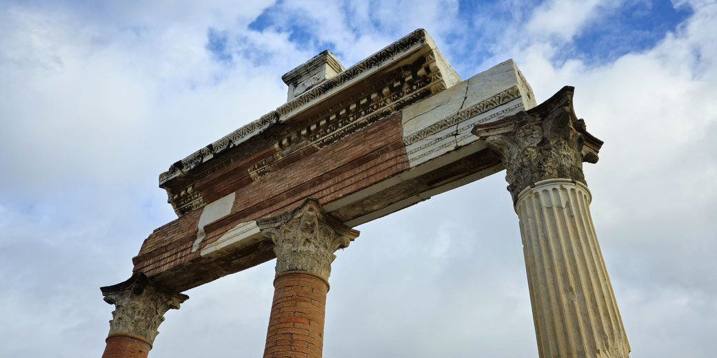 The remains of a Roman structure in Pompeii, featuring three weathered columns with ornate capitals supporting a partially intact entablature. The sky is cloudy in the background.