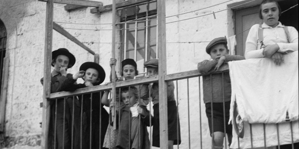 A group of children stand on a balcony behind a railing, gazing outward with somber expressions, some leaning on the bars.