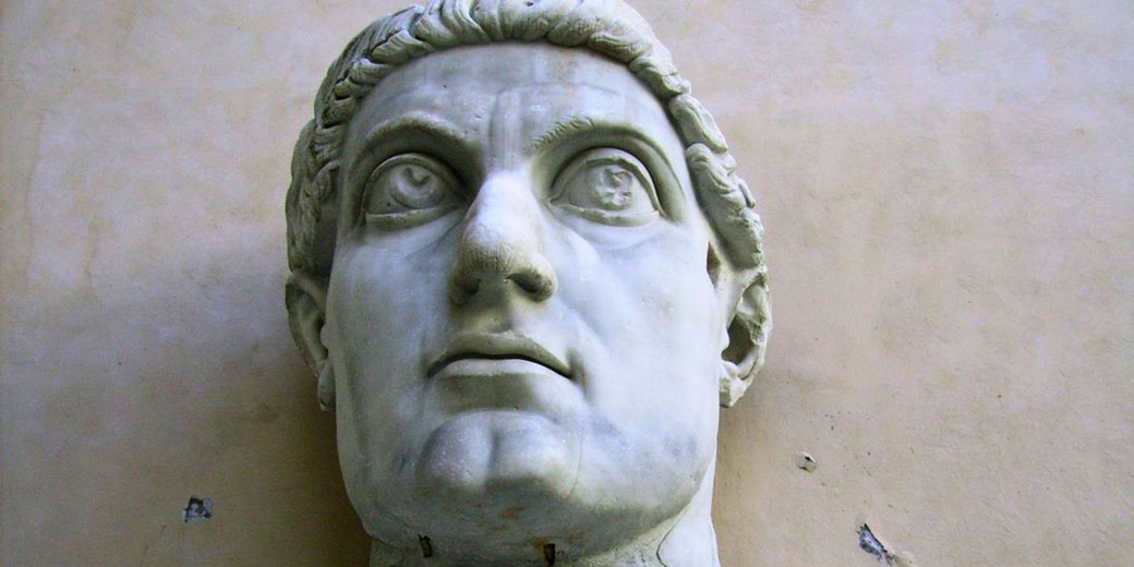Giant marble head of a Roman figure with wide eyes and detailed curls, mounted against a plain wall.