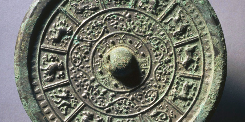 An ancient Chinese bronze mirror with intricate interlacing floral motifs and twelve zodiac animals. A raised central knob is surrounded by detailed carvings.