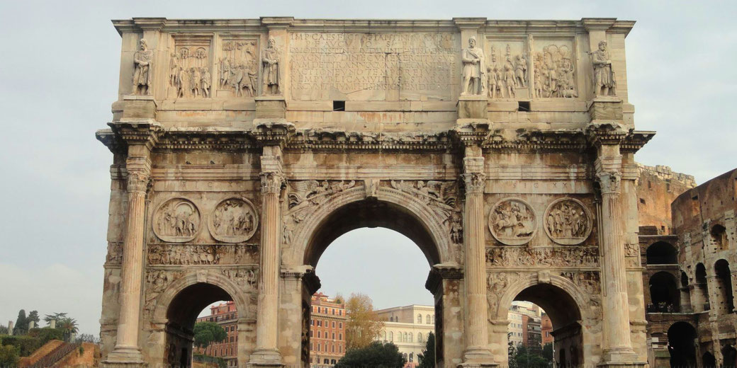 Ancient Roman arch with detailed reliefs and inscriptions, set against a backdrop of historic buildings and the Colosseum under an overcast sky.