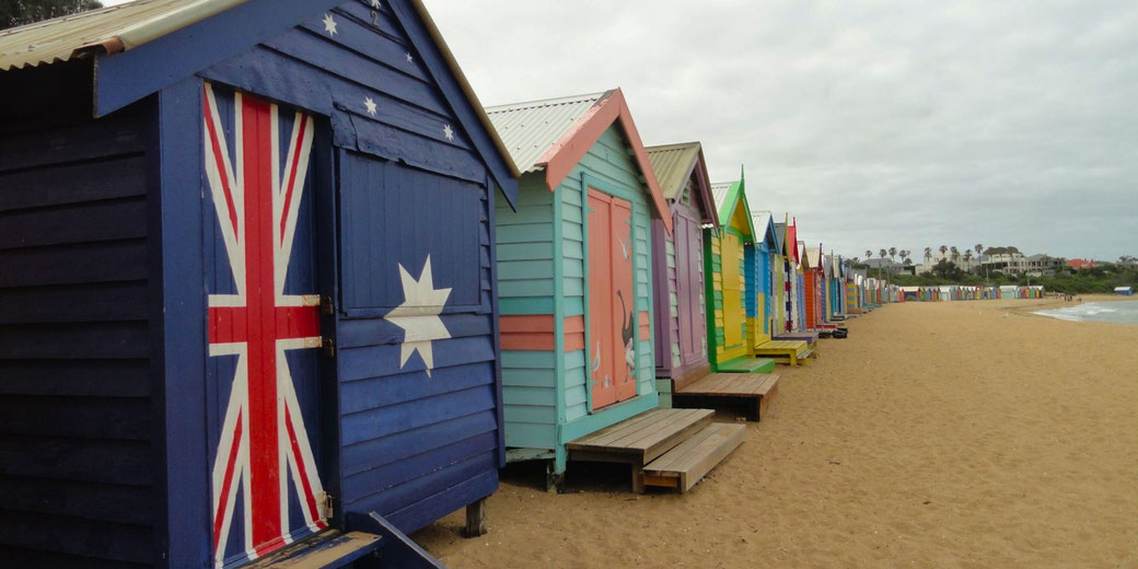 Colorful beach huts on a sandy shore, with the first hut painted like the Australian flag. The huts extend into the distance, with the ocean and palm trees visible under a cloudy sky.