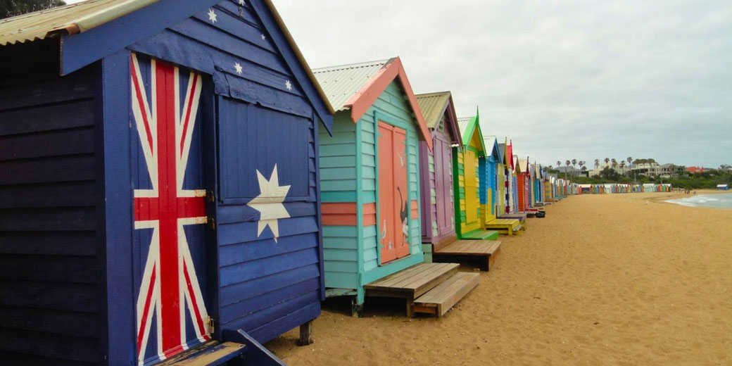 Colorful beach huts on a sandy shore, with the first hut painted like the Australian flag. The huts extend into the distance, with the ocean and palm trees visible under a cloudy sky.