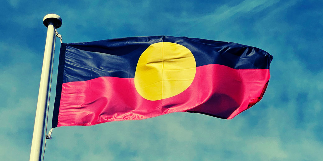 The Aboriginal flag waves on a pole with a blue sky backdrop, yellow circle centered on a black and red horizontal split.