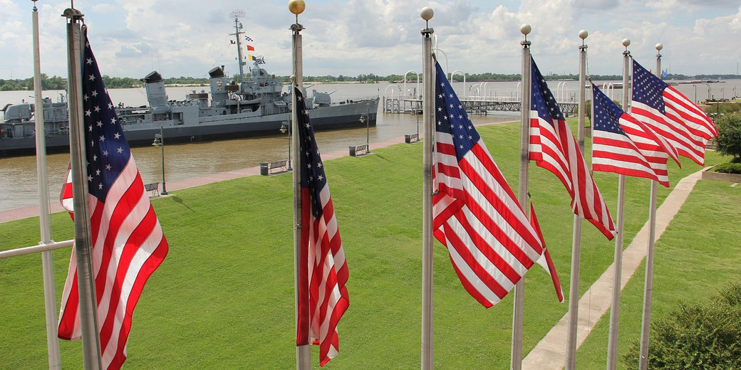 Multiple American flags line a walkway near a river with a gray naval ship docked in the background.