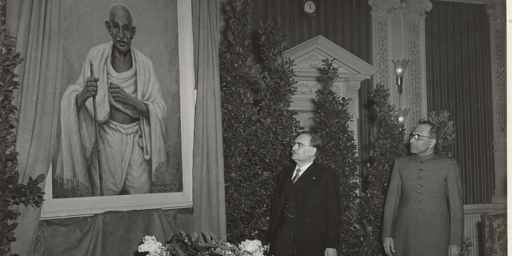 Two men stand beside a large portrait of Mahatma Gandhi during an unveiling ceremony, surrounded by plants and drapery.