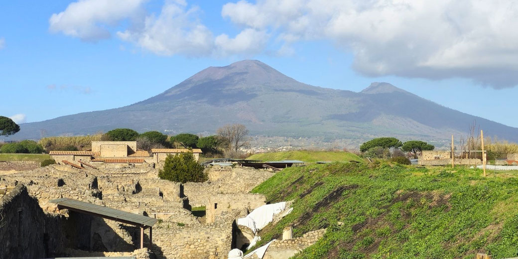Eruption of Vesuvius hour by hourRuins of an ancient city lie in the foreground with Mount Vesuvius towering in the background under a blue sky with scattered clouds.