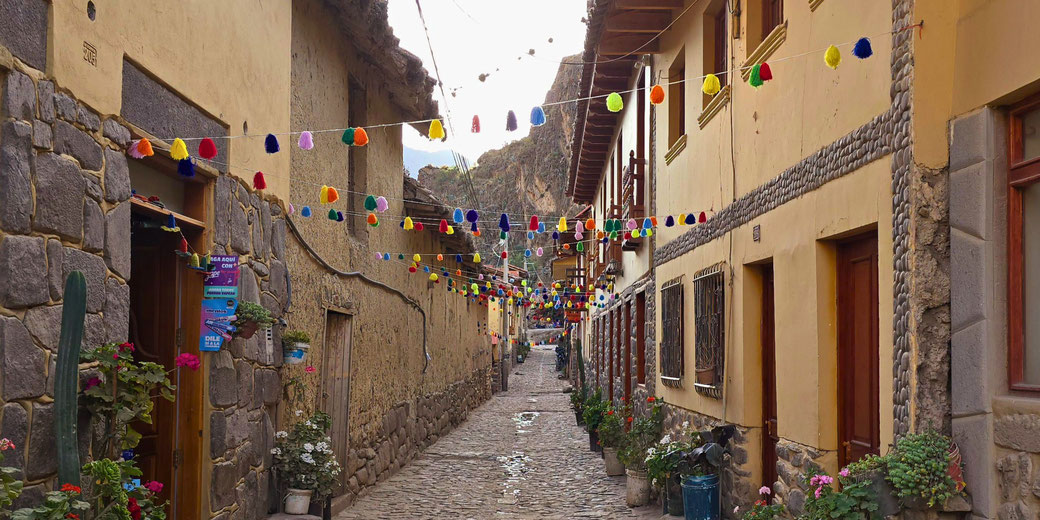 A charming cobblestone street in Ollantaytambo, Peru, adorned with colorful hanging tassels, rustic stone and adobe buildings, and lush potted plants, evoking a festive Andean atmosphere.