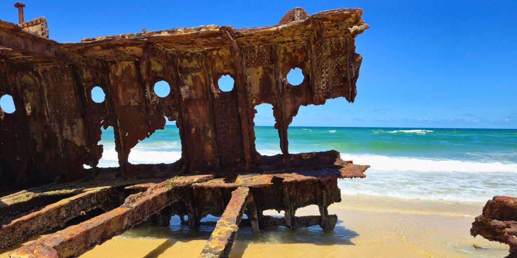 Close view of the S.S. Maheno wreck shows rusted steel hull panels with circular portholes, jagged edges, and exposed internal beams resting on damp sand as waves roll in behind.