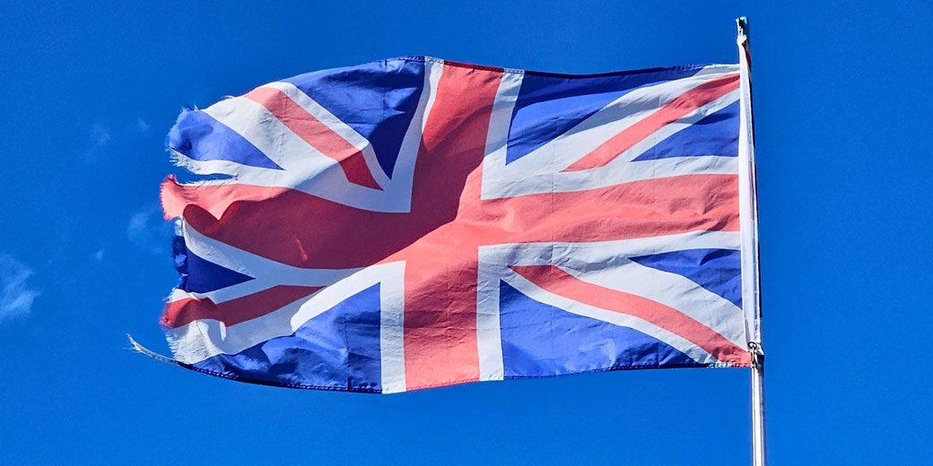 A worn Union Jack flag flutters in the wind against a clear blue sky on a sunny day.