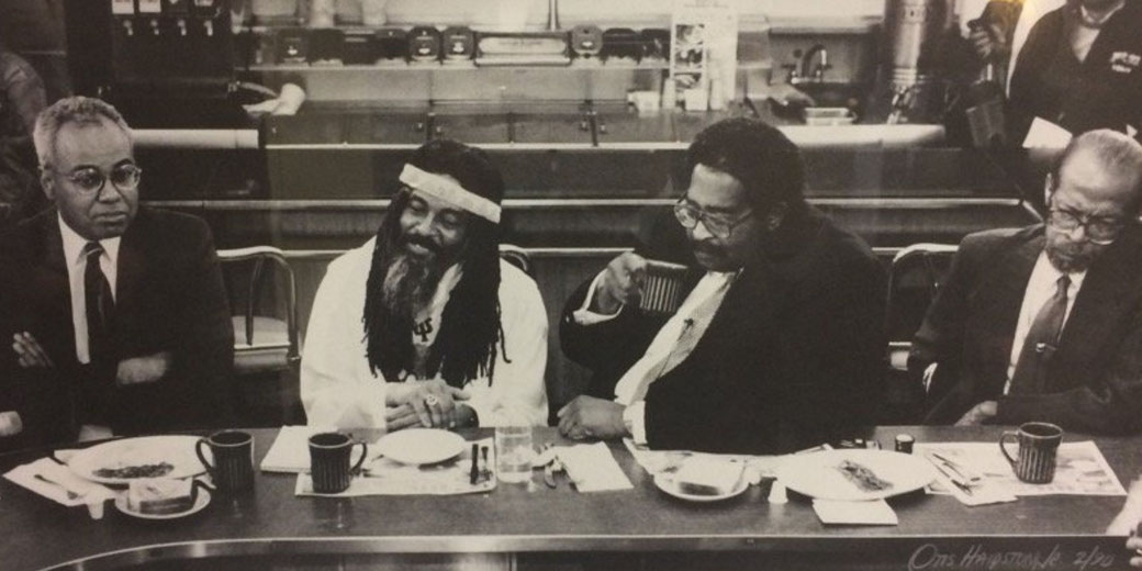 Four men sit at a diner counter, some eating and drinking, in a black-and-white photo capturing a casual yet reflective moment.