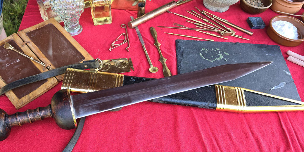 Roman-era artifacts on a red cloth, including a gladius sword, writing tablets, styluses, bronze tools, and small containers, suggesting a mix of military and daily life items.