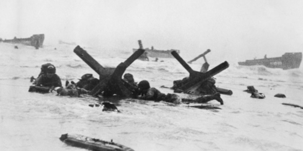 Soldiers crouch behind beach obstacles during a WWII amphibious assault under rough sea and weather conditions.