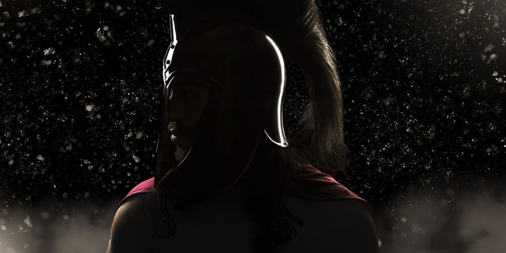 Silhouetted warrior in a plumed helmet looks sideways amid a dark, misty backdrop with scattered glowing particles.
