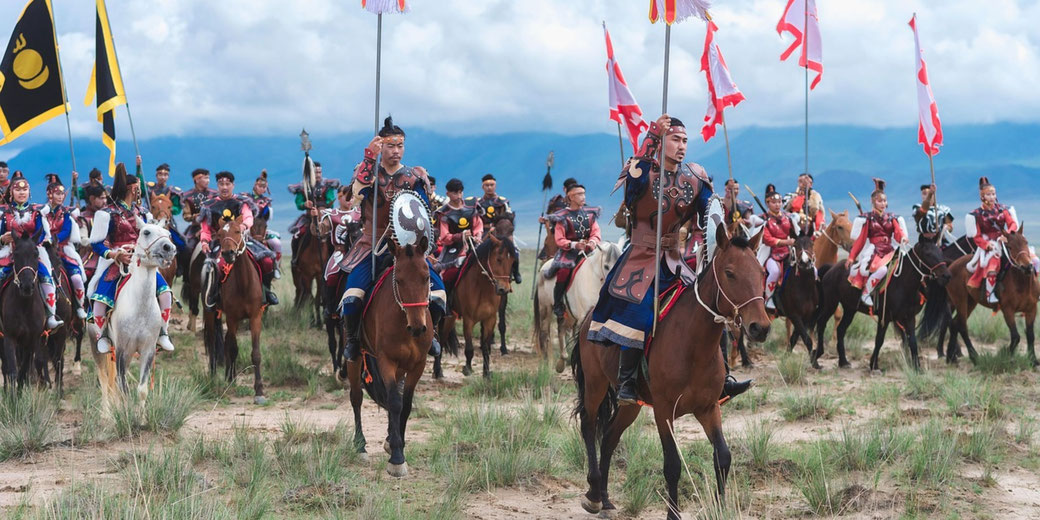 A group of riders in traditional Mongolian armor and colorful outfits march on horseback, carrying flags. 