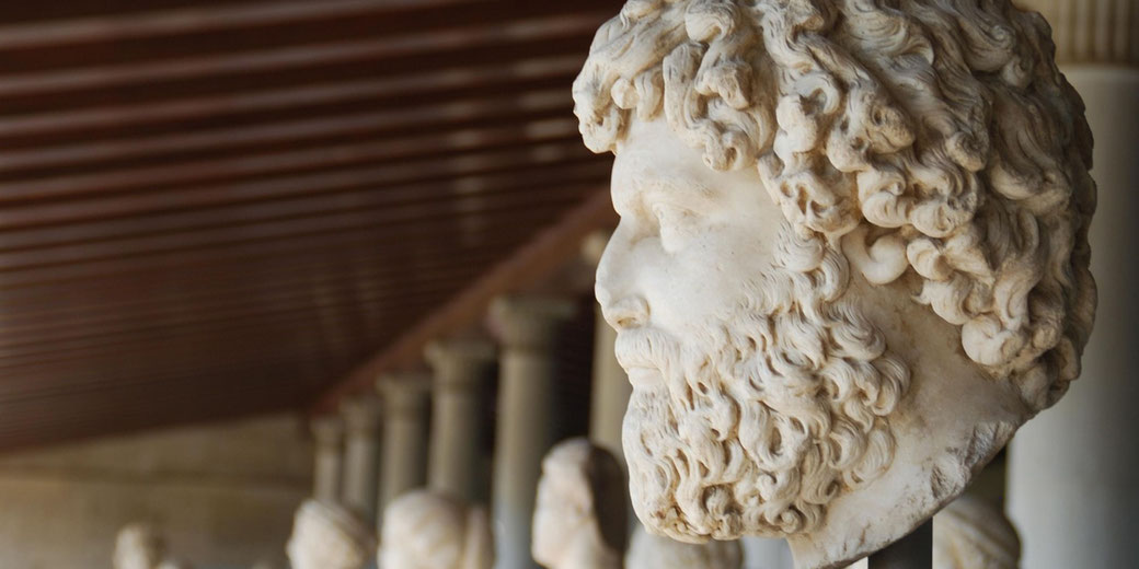 Marble bust of a bearded man with curly hair in profile, displayed among other sculptures in a colonnaded gallery.