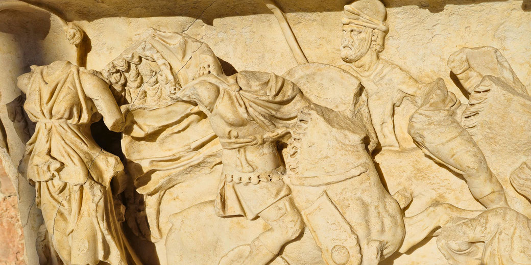 Ancient stone relief depicting a chaotic battle scene with warriors, horses, and flowing garments; some figures are headless or damaged.