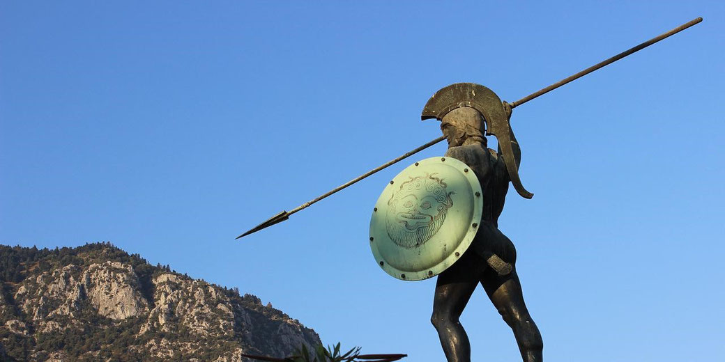Bronze statue of a Greek warrior holding a spear and shield, facing a rocky mountain under a clear blue sky.