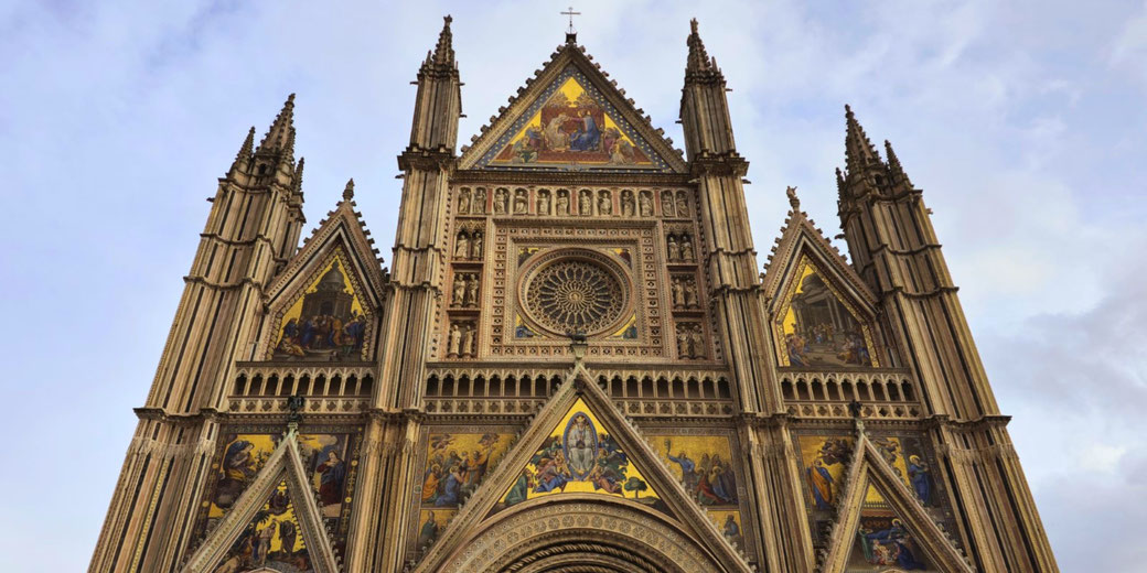 Gothic cathedral façade with tall spires, intricate carvings, and colorful religious mosaics under a cloudy sky.