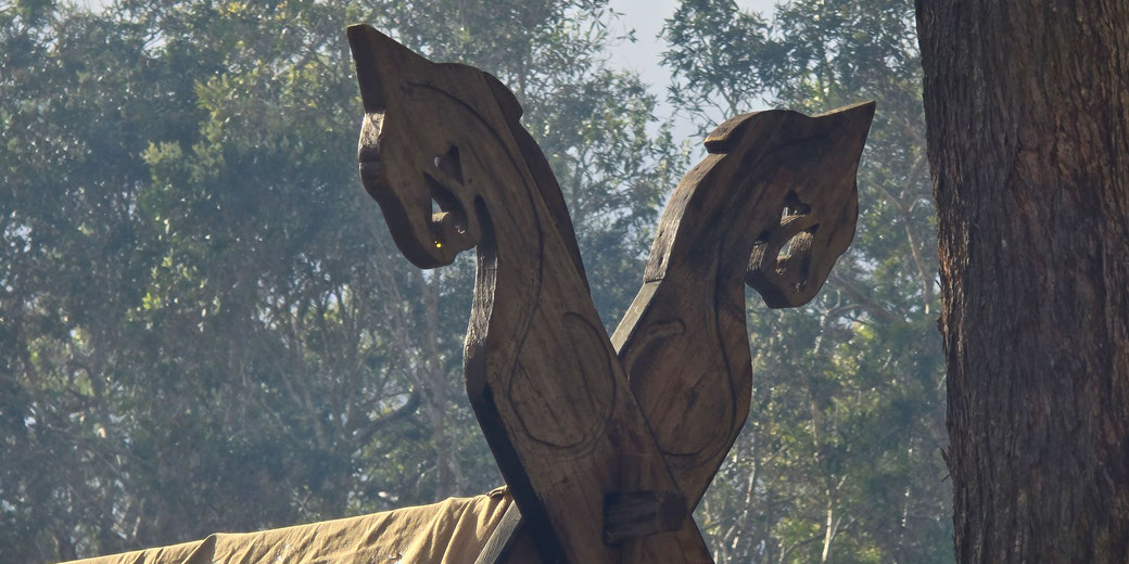 A close-up of intricately carved wooden dragon heads on the prow of a Viking-style longship, set against a forested background with soft light filtering through the trees.