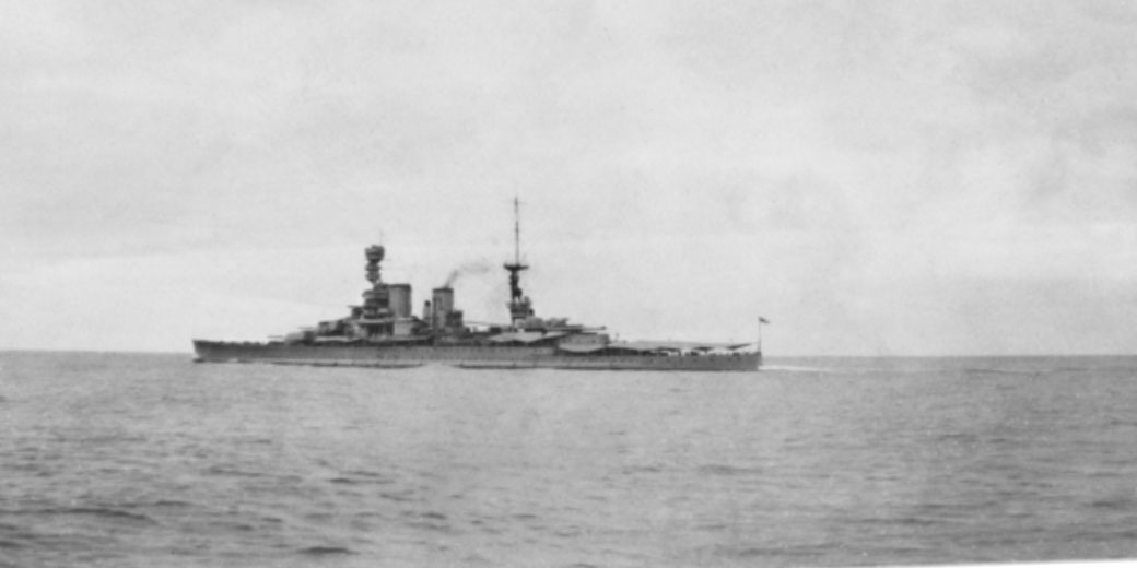 A large battleship sails across calm waters under a cloudy sky, viewed from a distance.