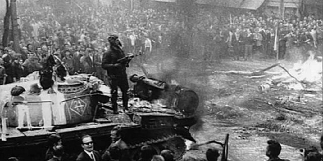 A Soviet tank burns in a Prague street during the August 1968 invasion, directly illustrating armed intervention and Czech civilian resistance.